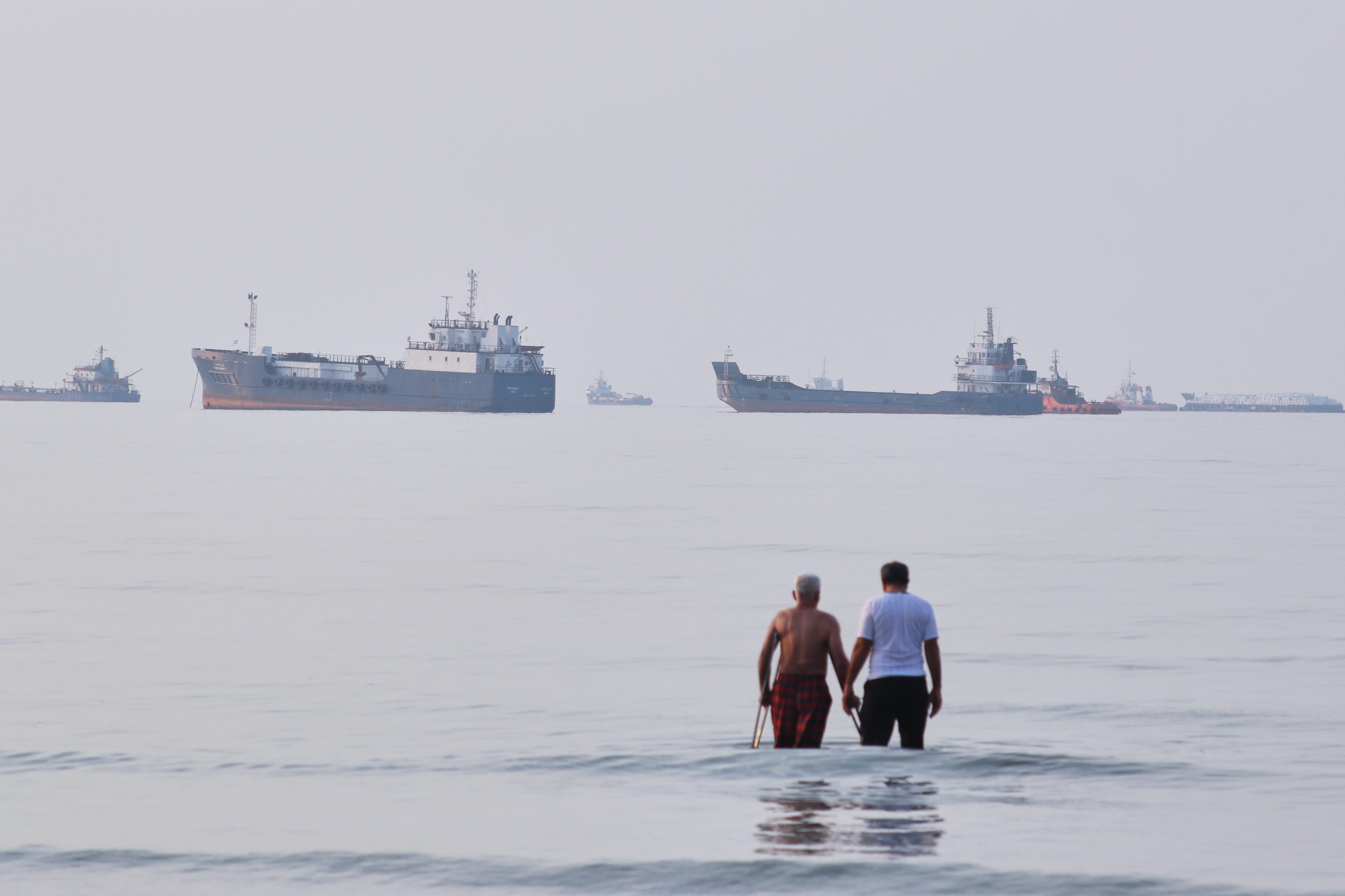 Two men wade in the waters of the Strait of Hormuz with vessels anchored in the background, off Bandar Abbas, Iran, Sunday, July 12, 2026.