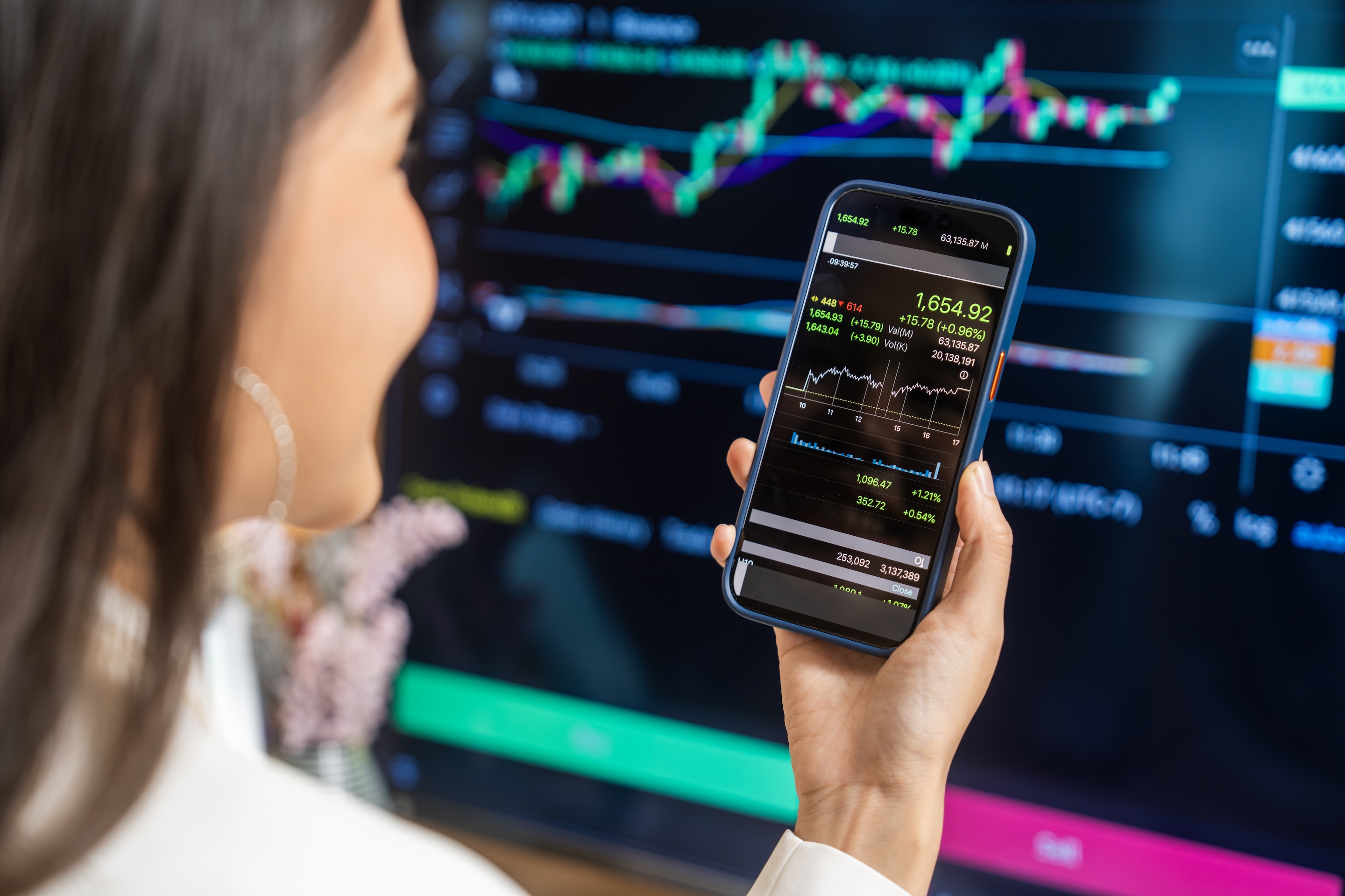 A woman observes stock trends on her smartphone while a computer screen displays financial graphs.
