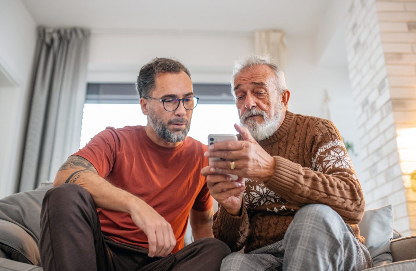 A younger man and an older man sit next to each other on a couch, looking down at a cell phone.