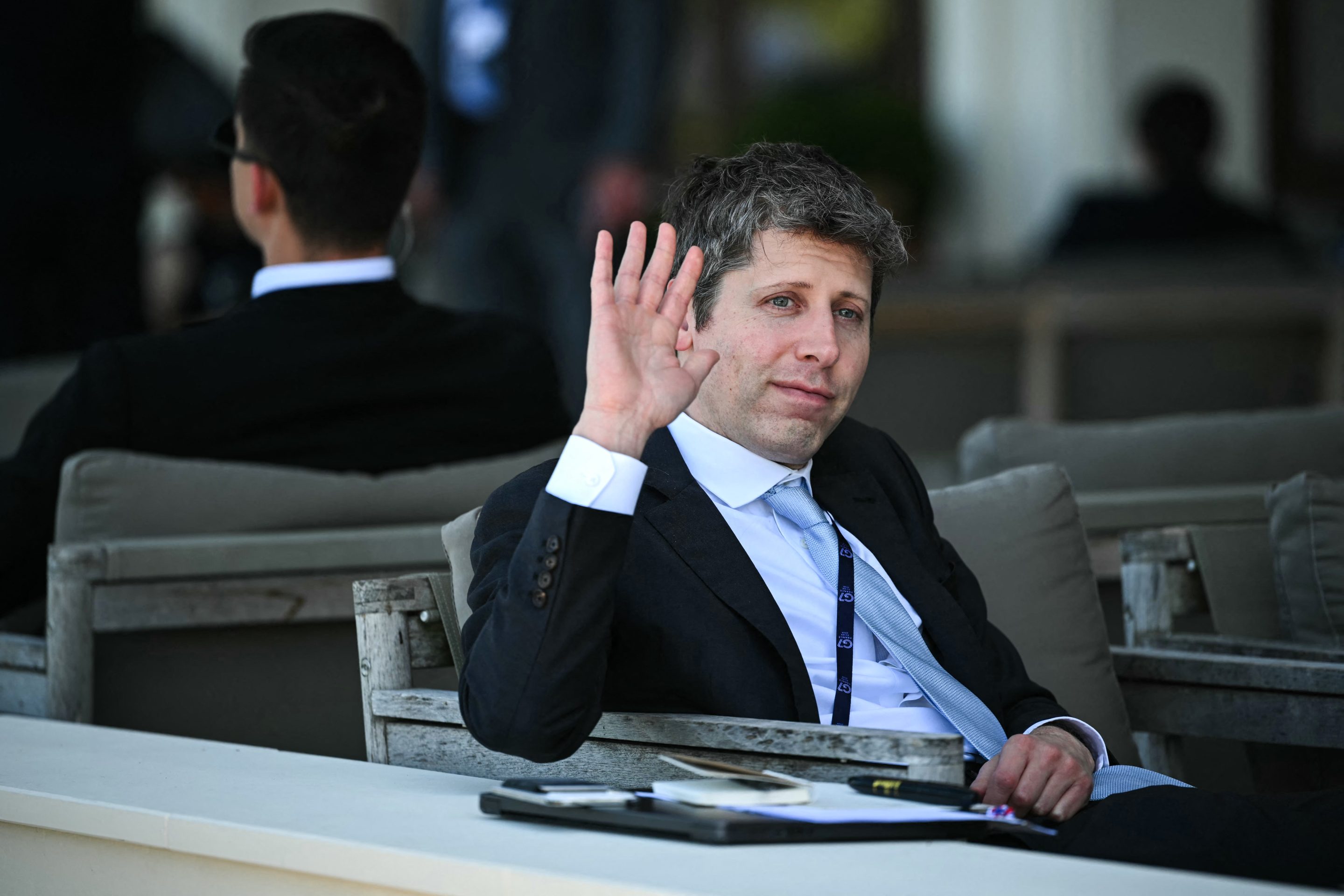 OpenAI Sam Altman sitting on a chair waving.